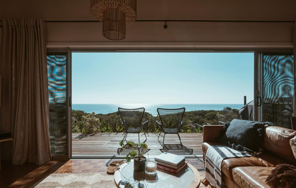 Cosy living room with ocean view, featuring two chairs on a deck, books, and a plant on a coffee table.