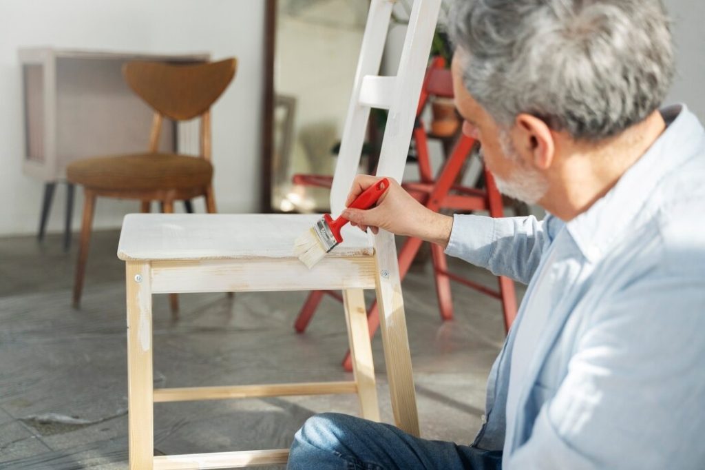 A man paints a wooden chair using a red paintbrush, focusing intently on his work