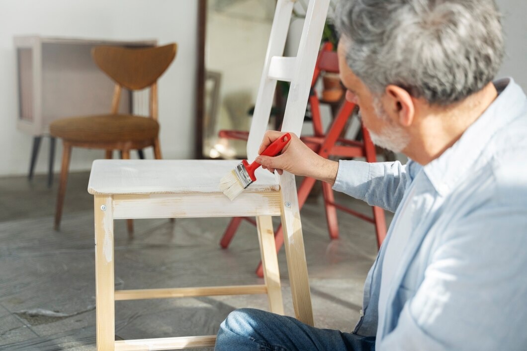 A man paints a wooden chair using a red paintbrush, focusing intently on his work